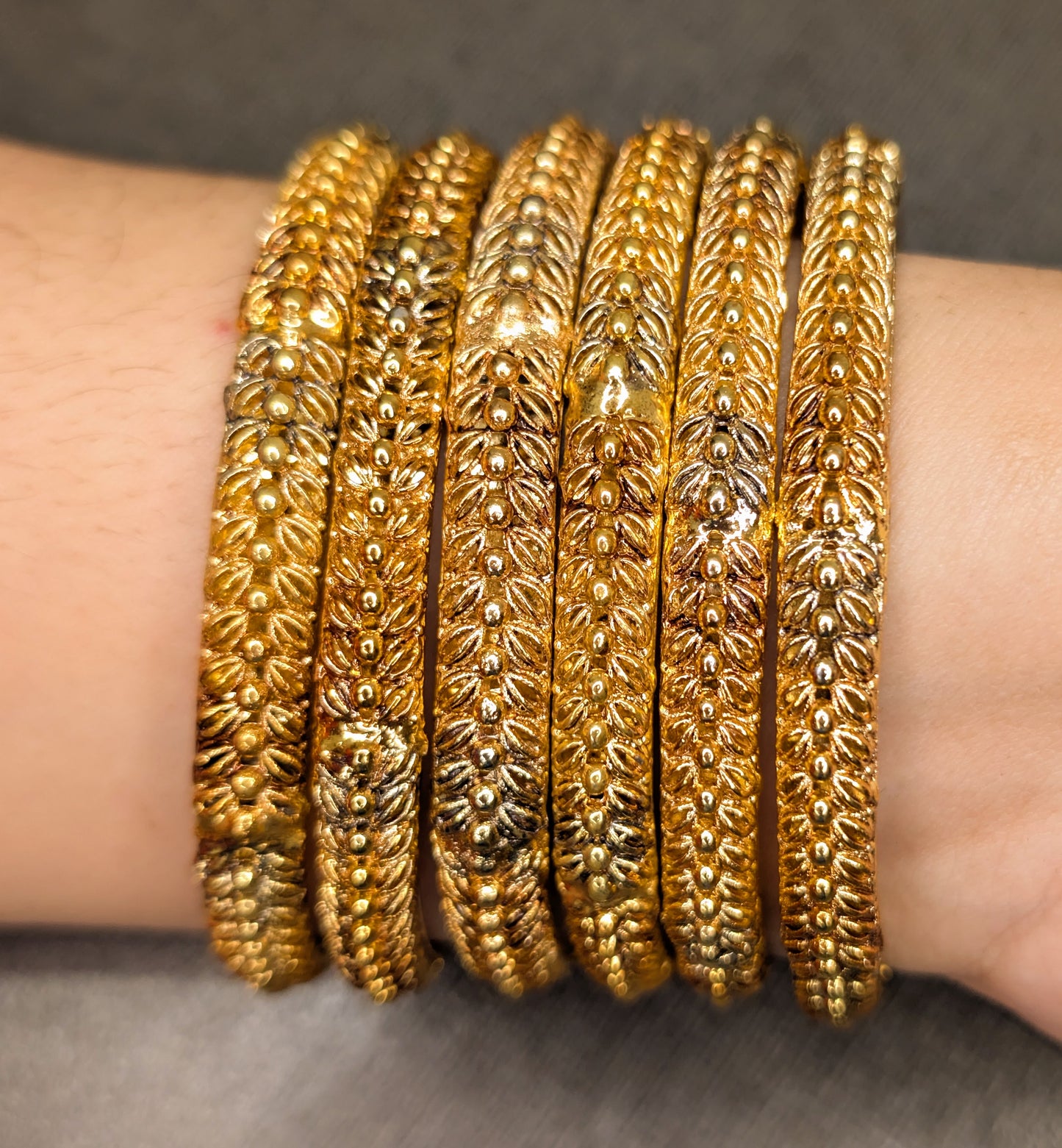 Close-up of a wrist wearing six gold bangles with intricate designs on a gray background
