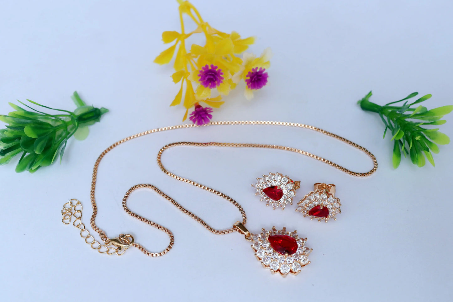 Gold necklace and earrings with red stones on a light blue background with decorative plants.