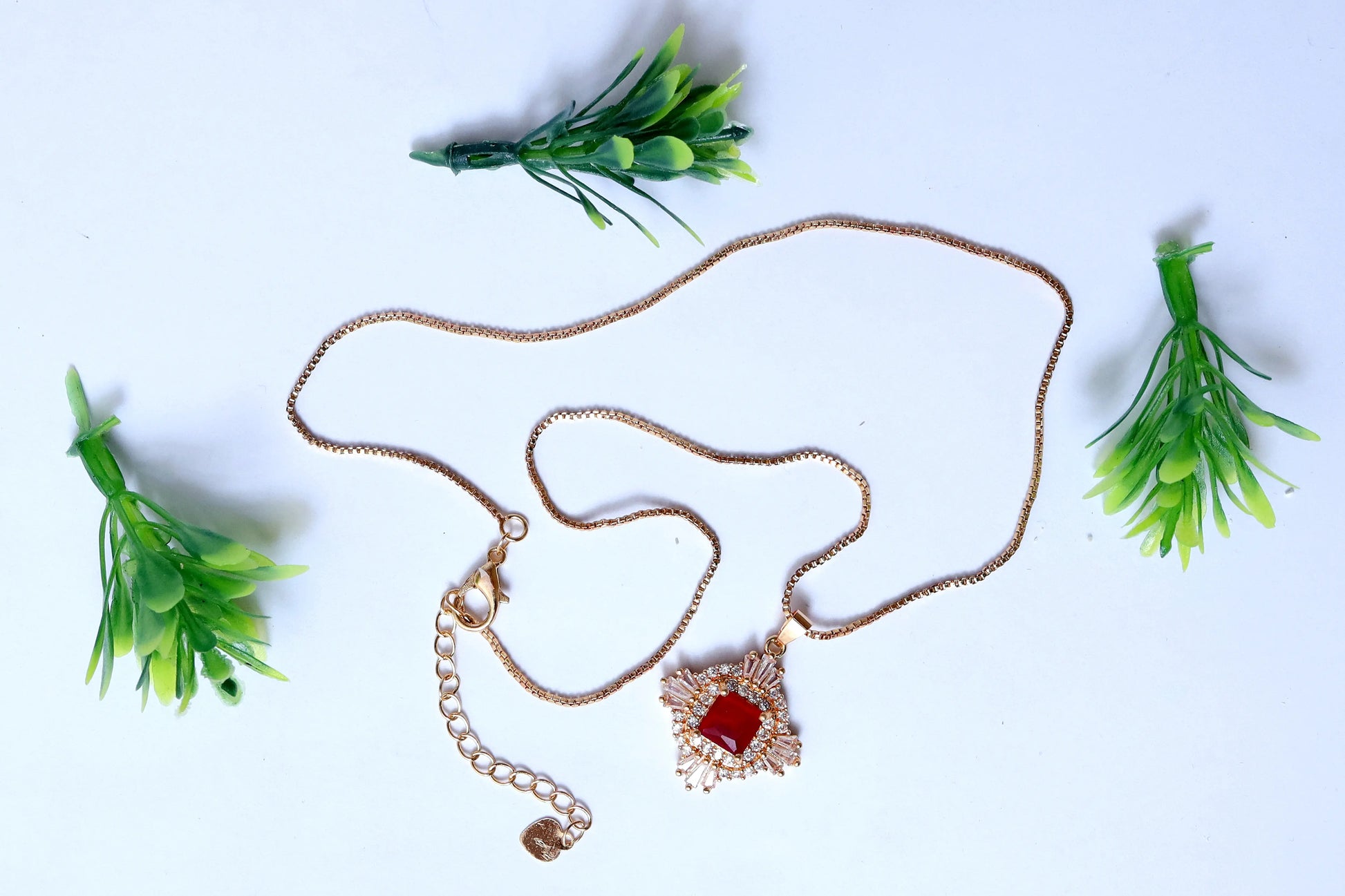 Gold necklace with a red pendant on a light blue background with green foliage.