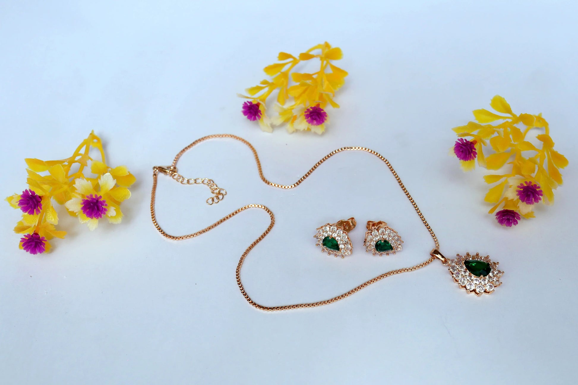 Gold necklace with heart pendant and matching earrings on a light blue background with yellow and pink flowers.