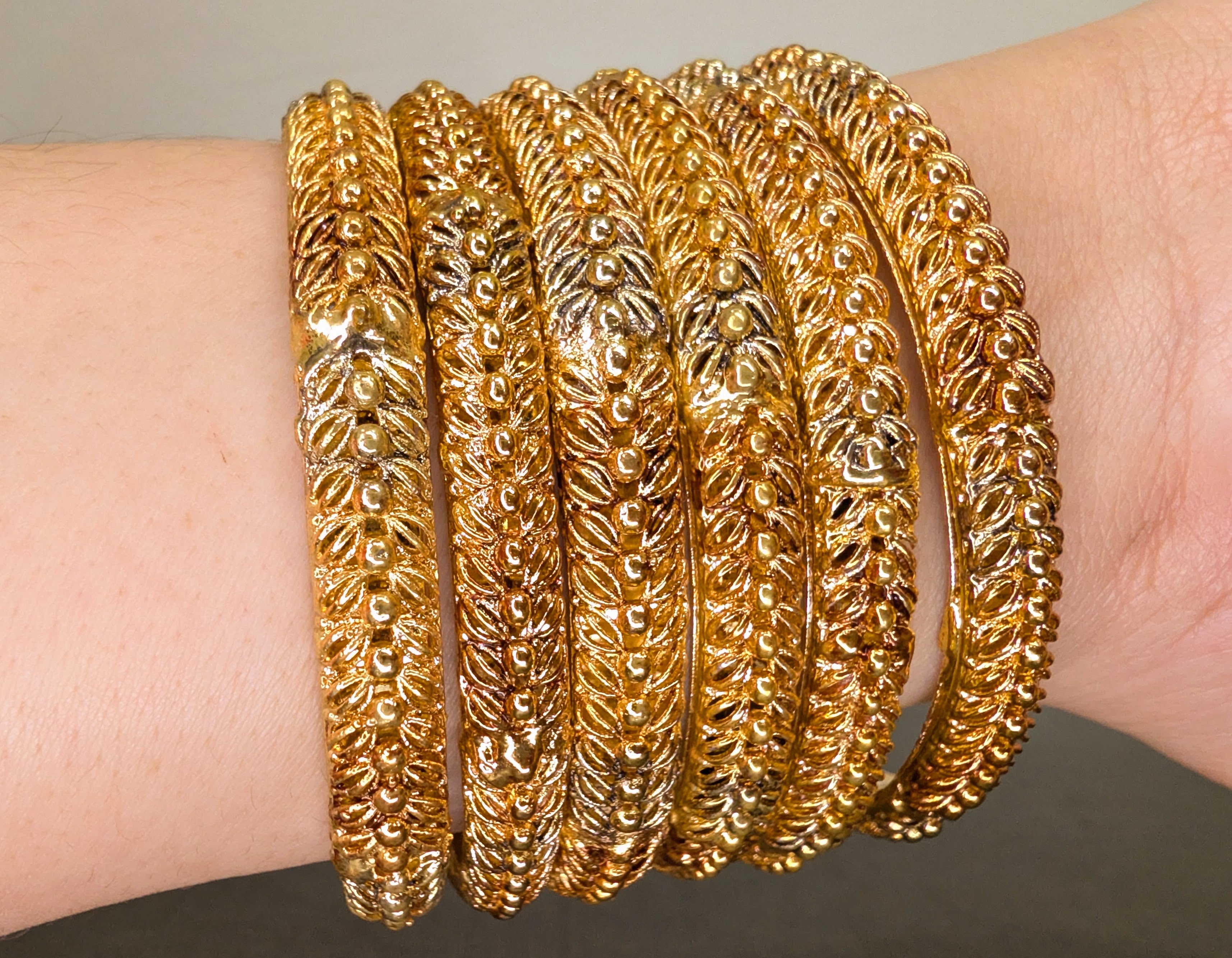 Gold bangles with intricate designs on a wrist against a neutral background