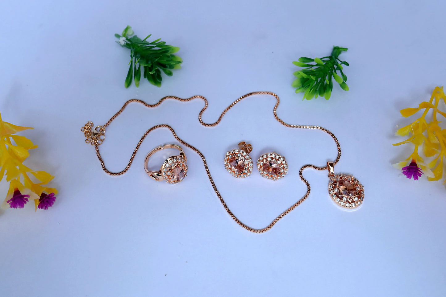Rose gold jewelry set with necklace, ring, and earrings on a light blue background with decorative plants and flowers.