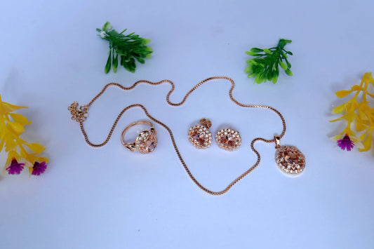 Rose gold jewelry set with necklace, ring, and earrings on a light blue background with decorative plants and flowers.