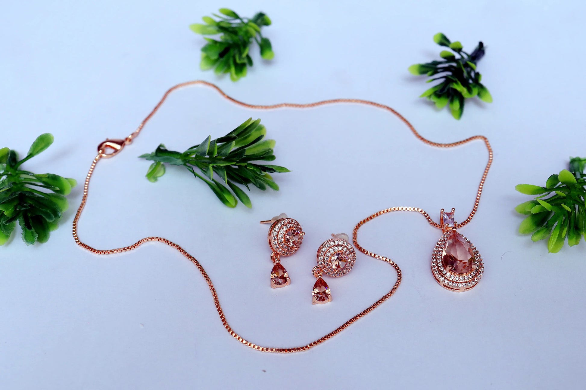 Rose gold jewelry set with necklace, earrings, and pendant on a light blue background with green plants.