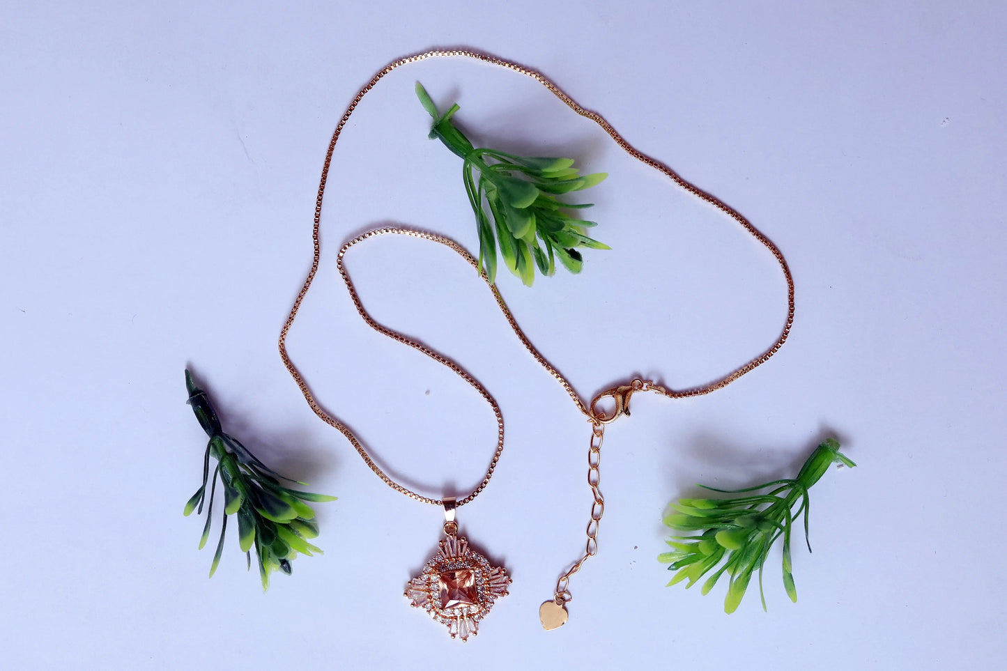 Rose gold necklace with a pendant on a light purple background with green leaves.