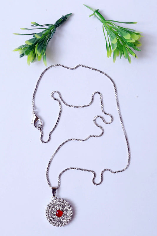 Silver necklace with a red gemstone pendant on a white background with green plants.