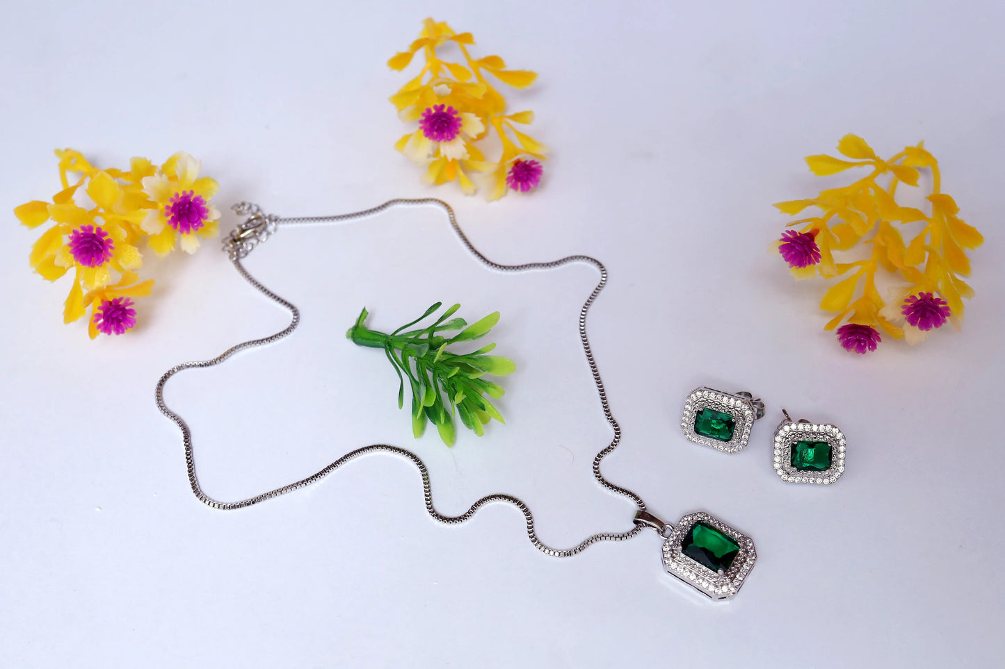 Silver necklace with green gemstones and matching earrings on a white background with yellow flowers.