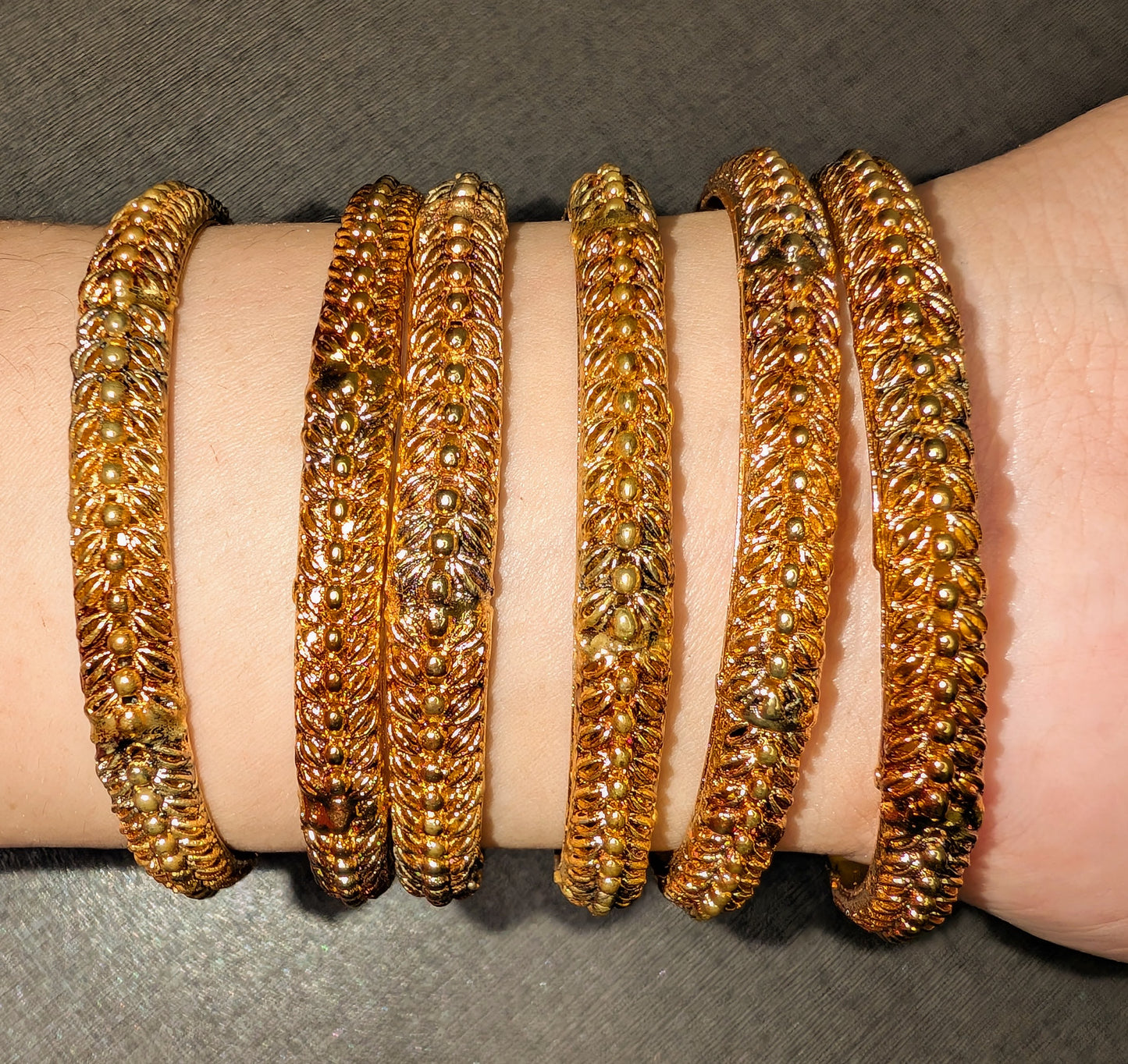 six gold bangles in gapes on hand or wrist with intricate designs on a wrist against a gray background
